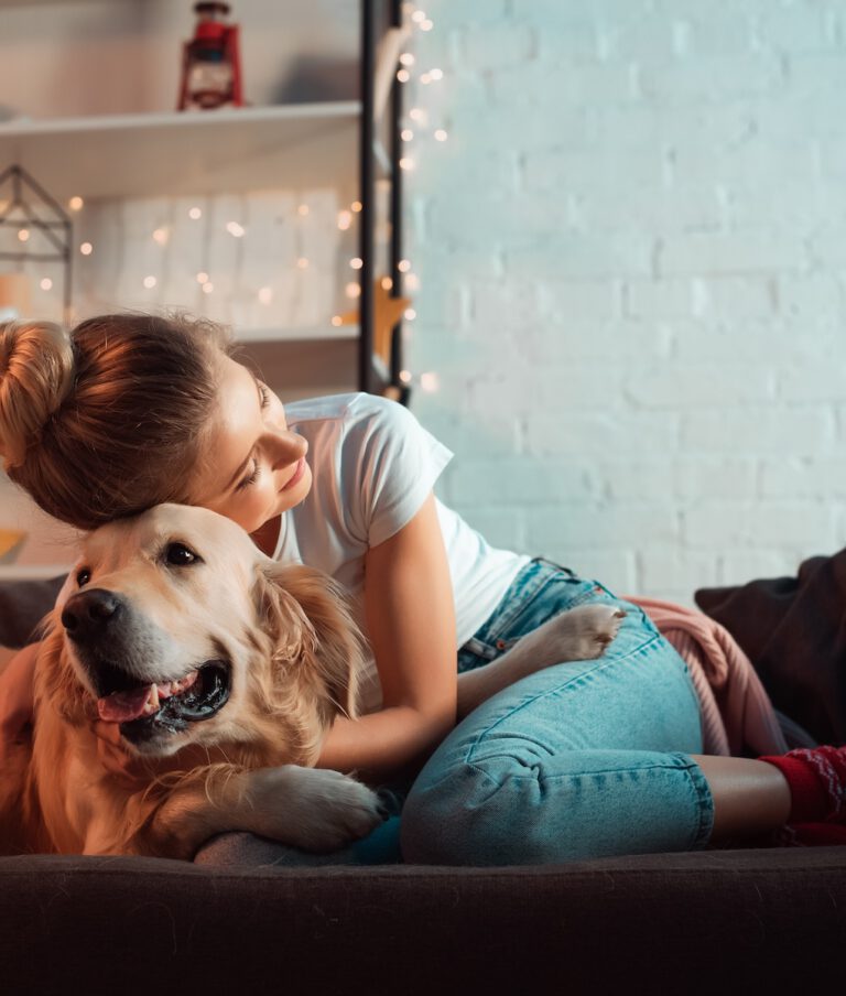 A women affectionately embraces a golden retriever on a couch. A cozy room setting with soft lighting and a white brick backdrop.